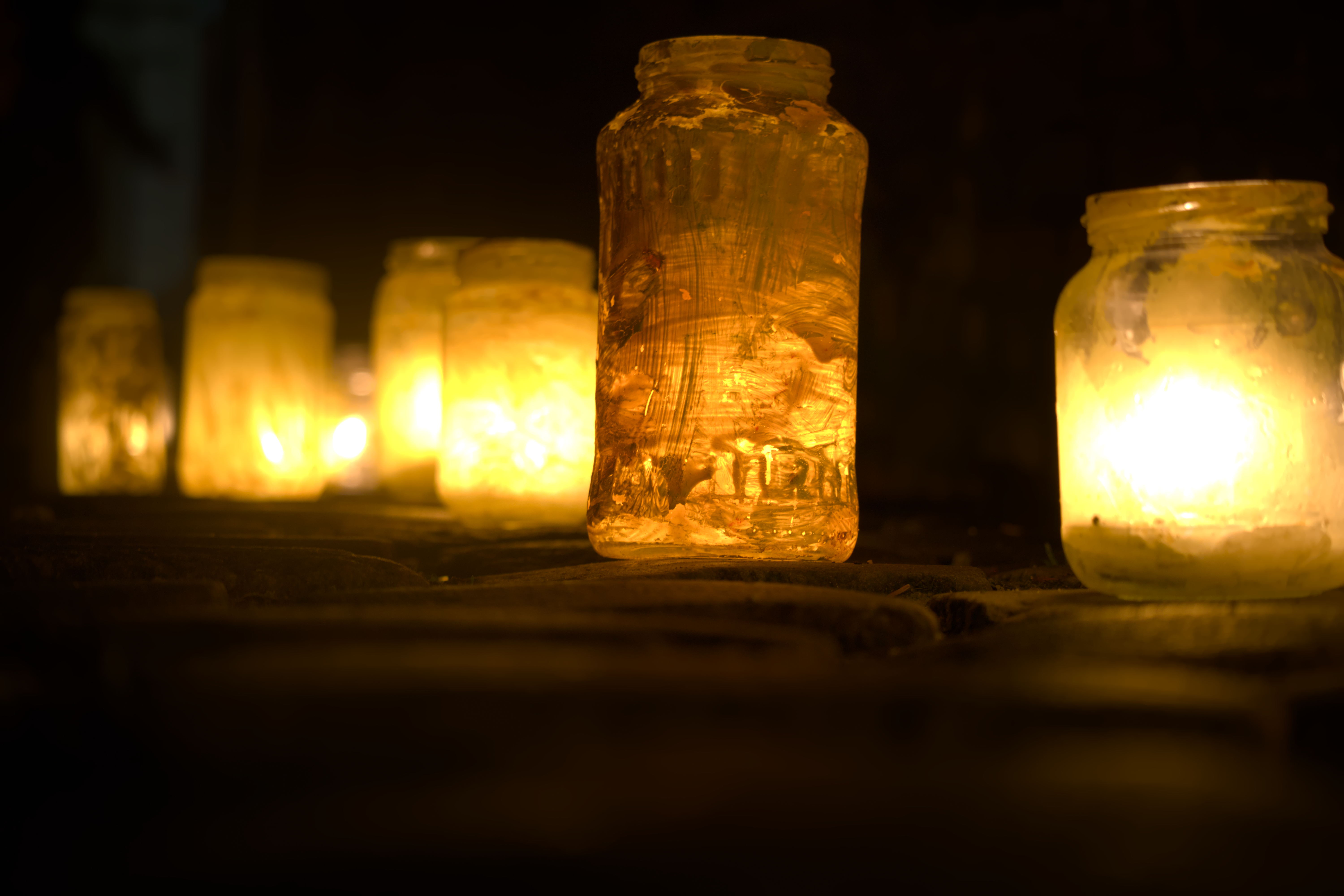 A orange/brownish painted glass, containing a candle, glowing onto a cobblestone pavement. More glasses in the bokeh.