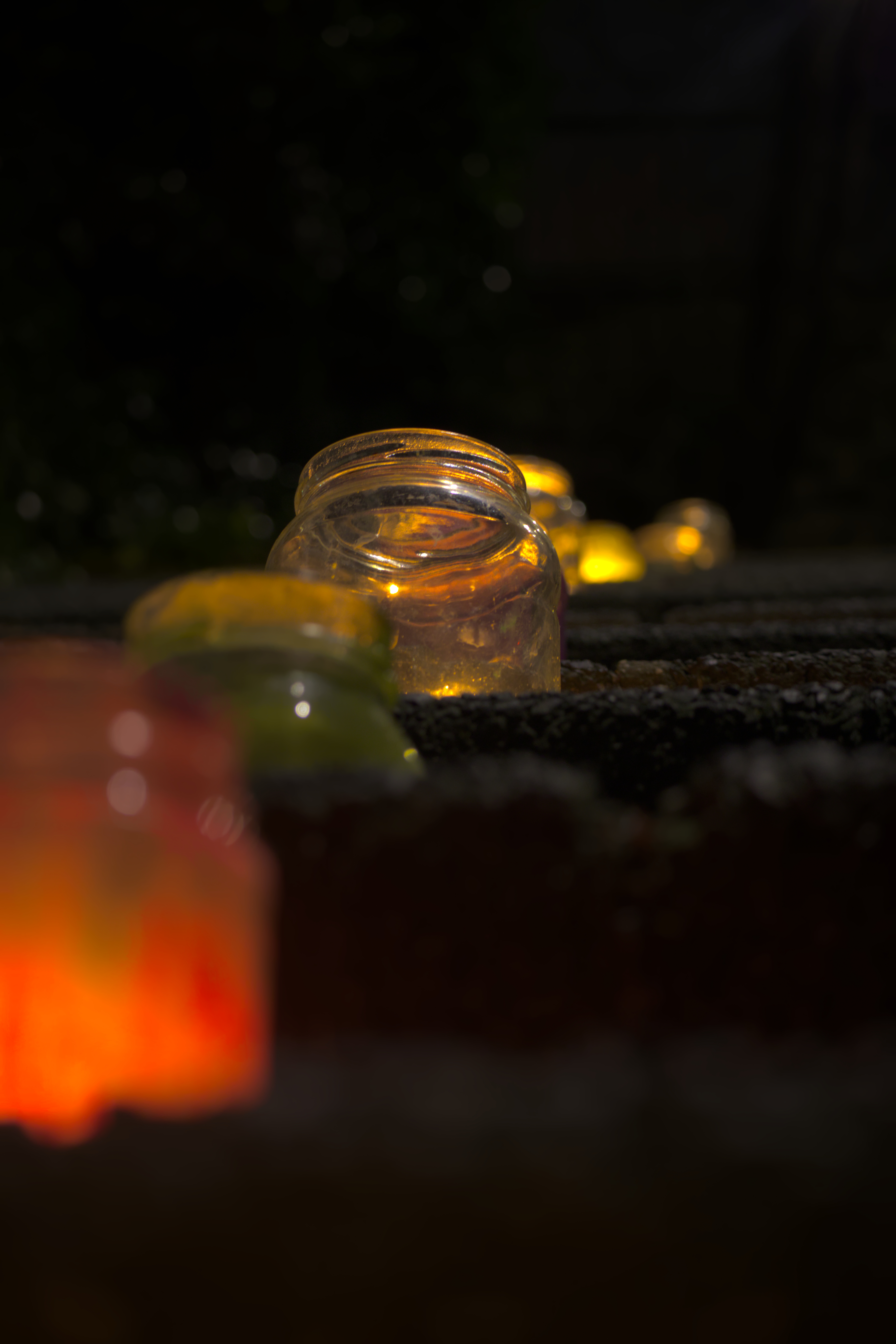 A row of colorfull glasses filled with candles lined up on stairs illuminting them. In focus is a transparent glass.